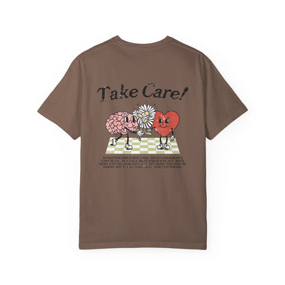Graphic tee featuring a whimsical design with a heart and brain holding a flower, paired with motivational text encouraging self-care and positivity. Perfect for mental health awareness and retro-inspired casual style.