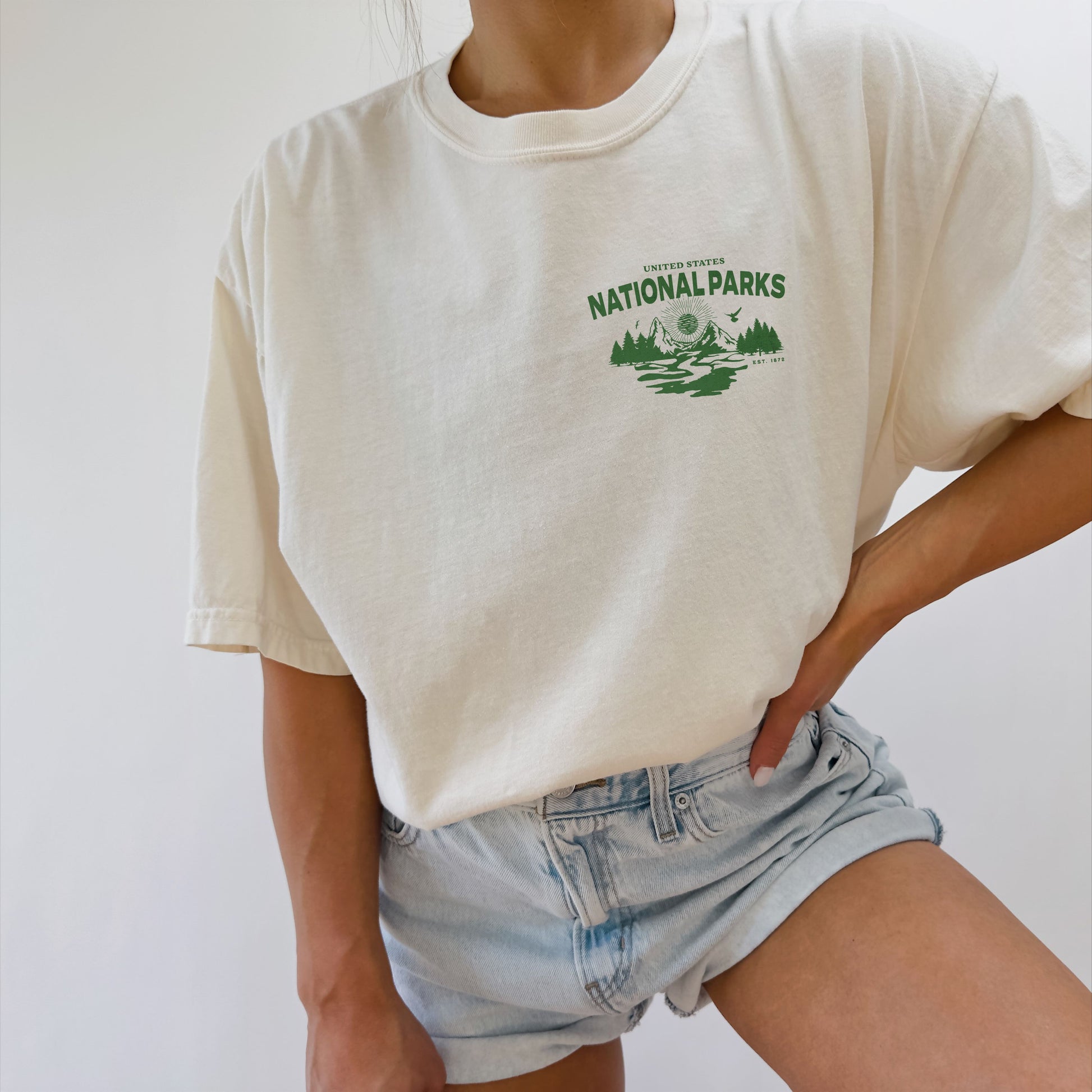 Person wearing a white t-shirt with 'National Parks' logo and denim shorts on a plain background