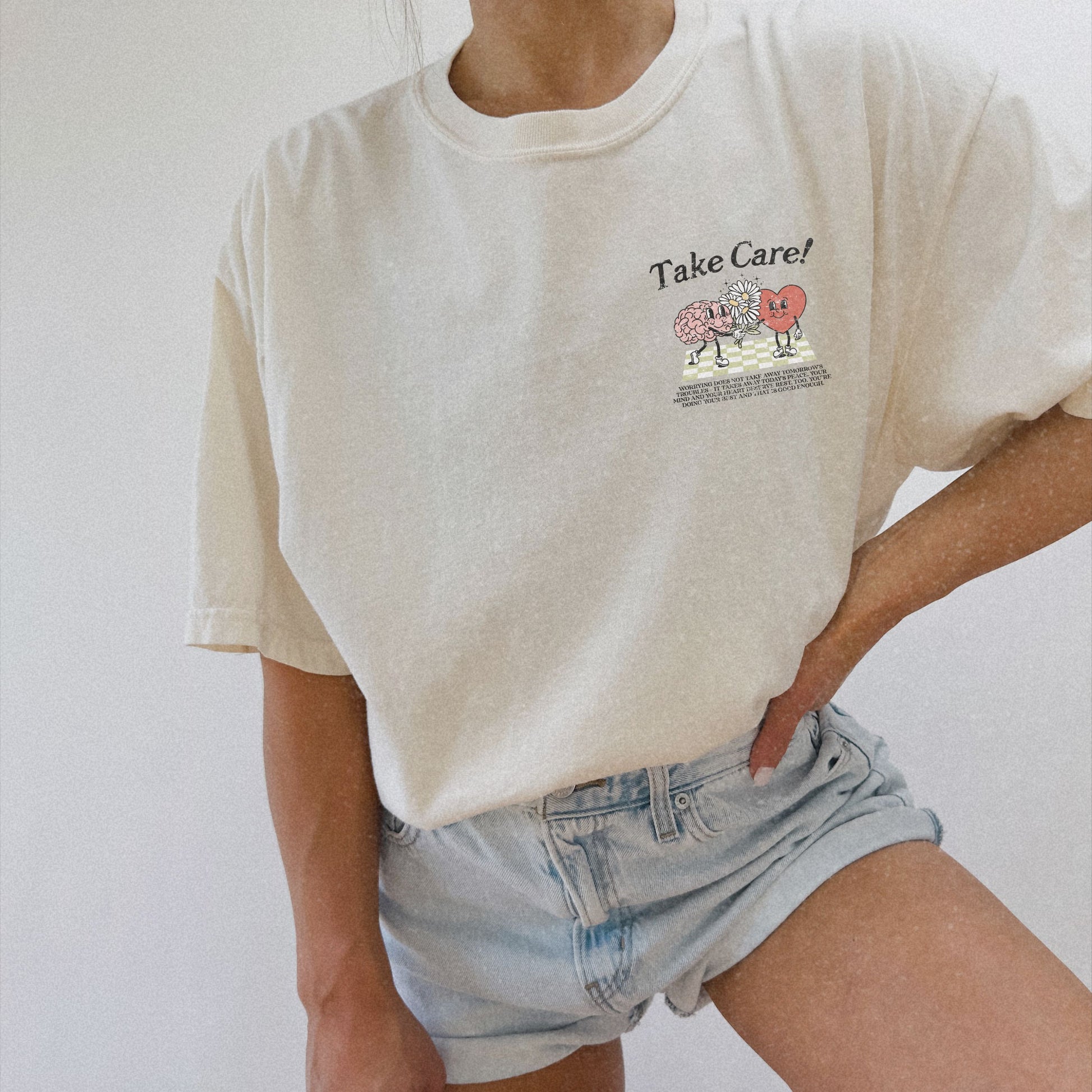 Graphic tee featuring a whimsical design with a heart and brain holding a flower, paired with motivational text encouraging self-care and positivity. Perfect for mental health awareness and retro-inspired casual style.