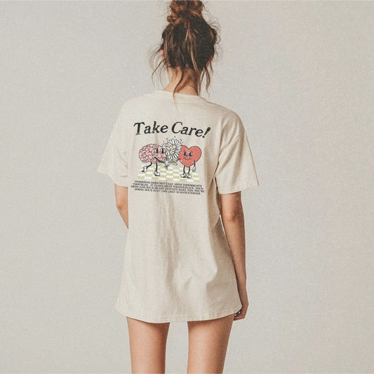 Graphic tee featuring a whimsical design with a heart and brain holding a flower, paired with motivational text encouraging self-care and positivity. Perfect for mental health awareness and retro-inspired casual style.